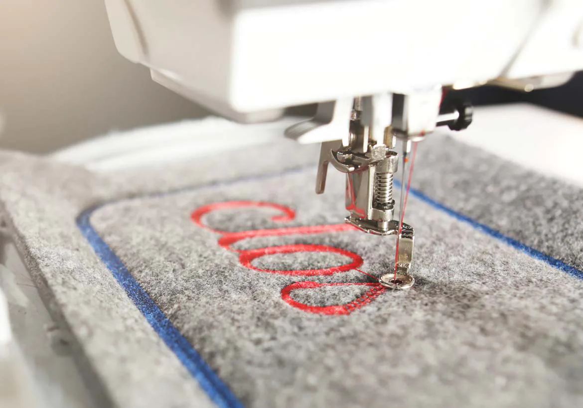 Close-up of embroidery machine stitching custom red lettering on fabric, showcasing precision in custom embroidery and apparel decoration – Custom Apparel, Screenprinting, and Embroidery services in Los Angeles.