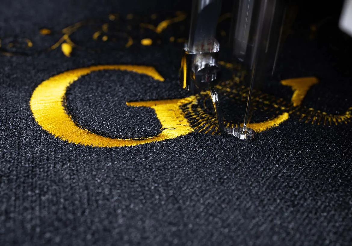 Close-up of embroidery machine stitching gold lettering on fabric, highlighting intricate and special embroidery techniques – Custom Apparel, Screenprinting, and Embroidery services in Los Angeles.